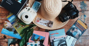 Flat lay of travel accessories including a straw hat, cameras, and guidebooks on a wooden table.